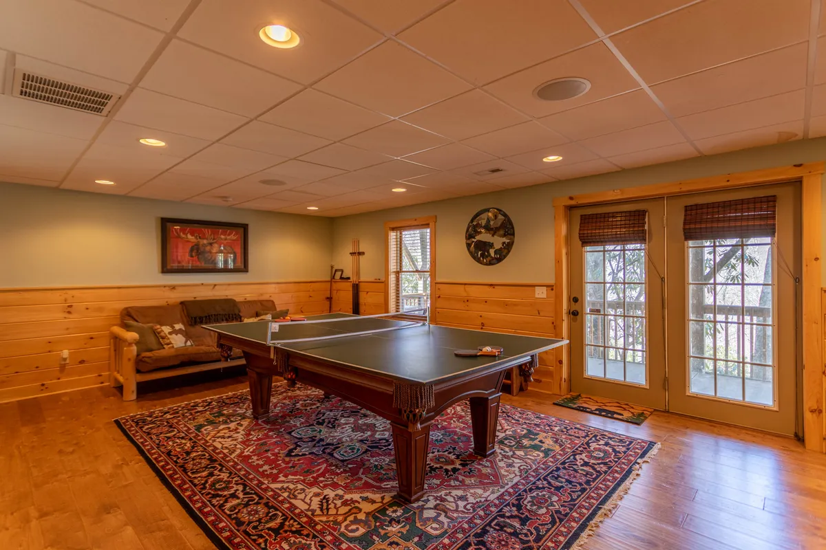 A spacious game room featuring a full-size pool table as the centerpiece, surrounded by a comfortable sofa and a plush rug. Double doors open out to a porch area, hinting at direct access to the outdoors. The warm wooden walls and relaxed seating create an inviting atmosphere for socializing and entertainment.