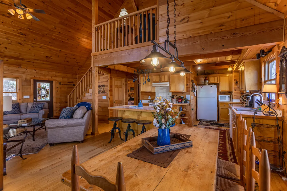 A panoramic shot of the main living area, featuring a wooden dining table, kitchen island, comfortable seating area, and large windows framing the surrounding woods. The space feels airy and connected, ideal for group meals, relaxation, and enjoying scenic views.