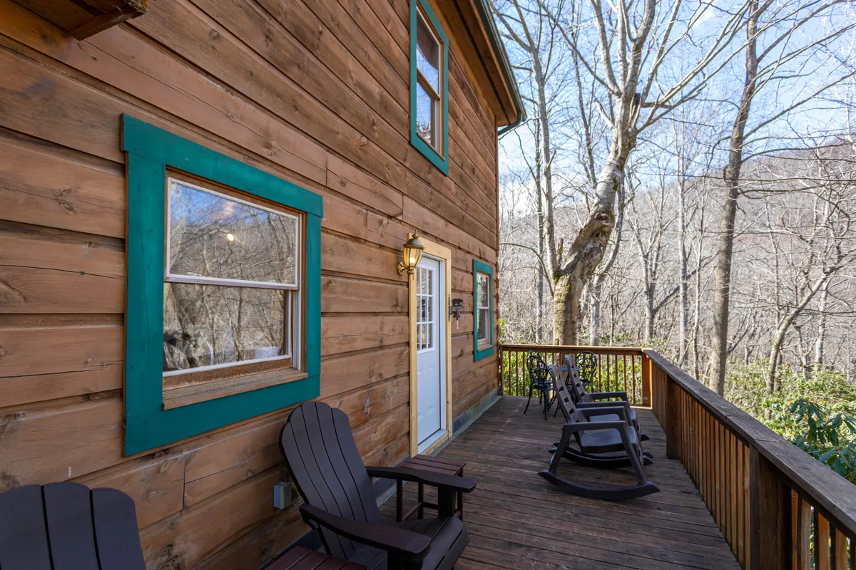 A rustic wooden deck with turquoise-trimmed windows and comfy rocking chairs provides a perfect spot for relaxation. The deck looks out onto serene nature, suggesting the ideal place to watch the sunrise or sunset, read a book, or enjoy conversation.