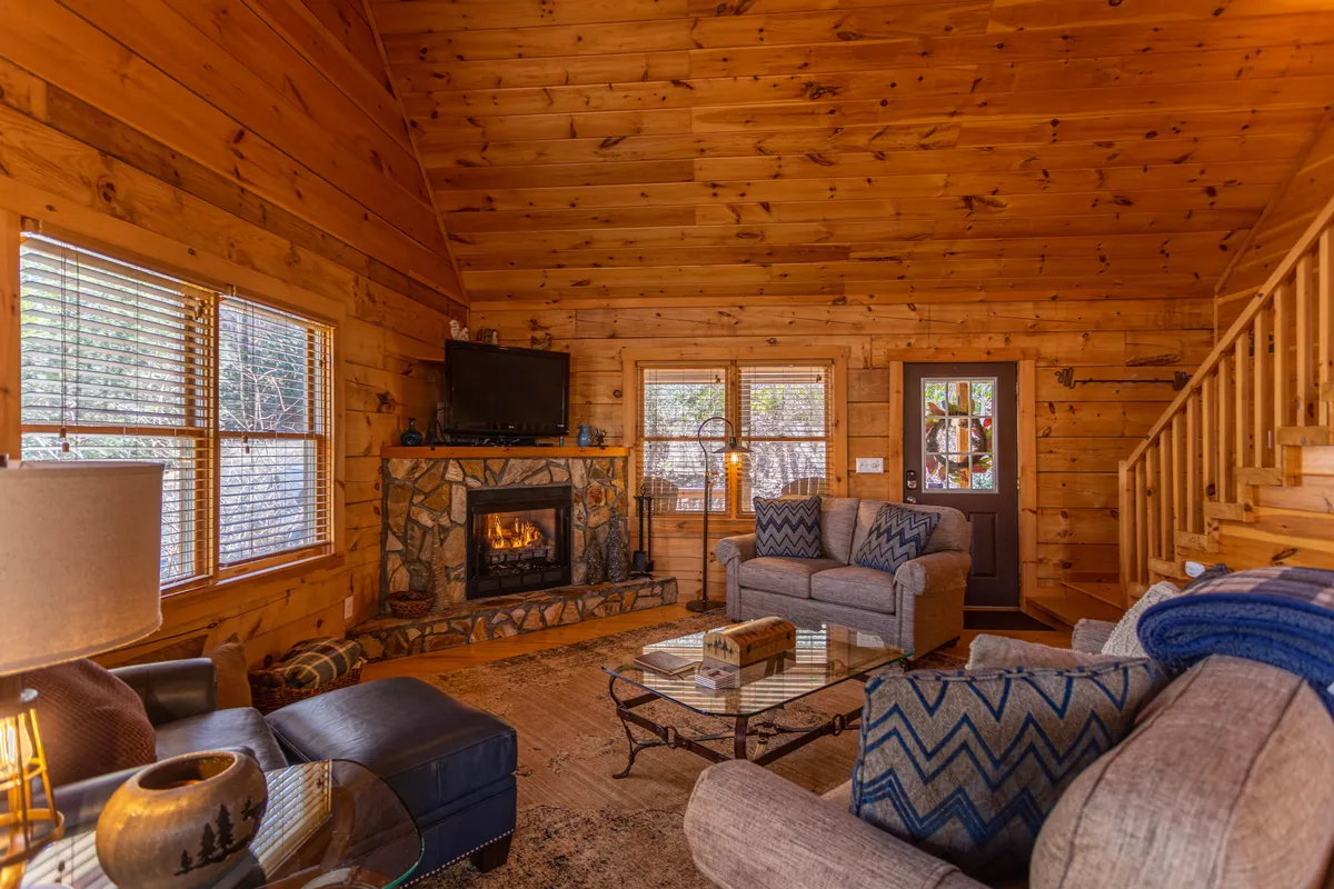 A cozy living area featuring a stone fireplace, plush sofa, and rustic wooden walls. Large windows let in soft natural light and offer glimpses of the tranquil woods outside. The coffee table is set for a relaxing morning, and the overall atmosphere is warm and inviting.