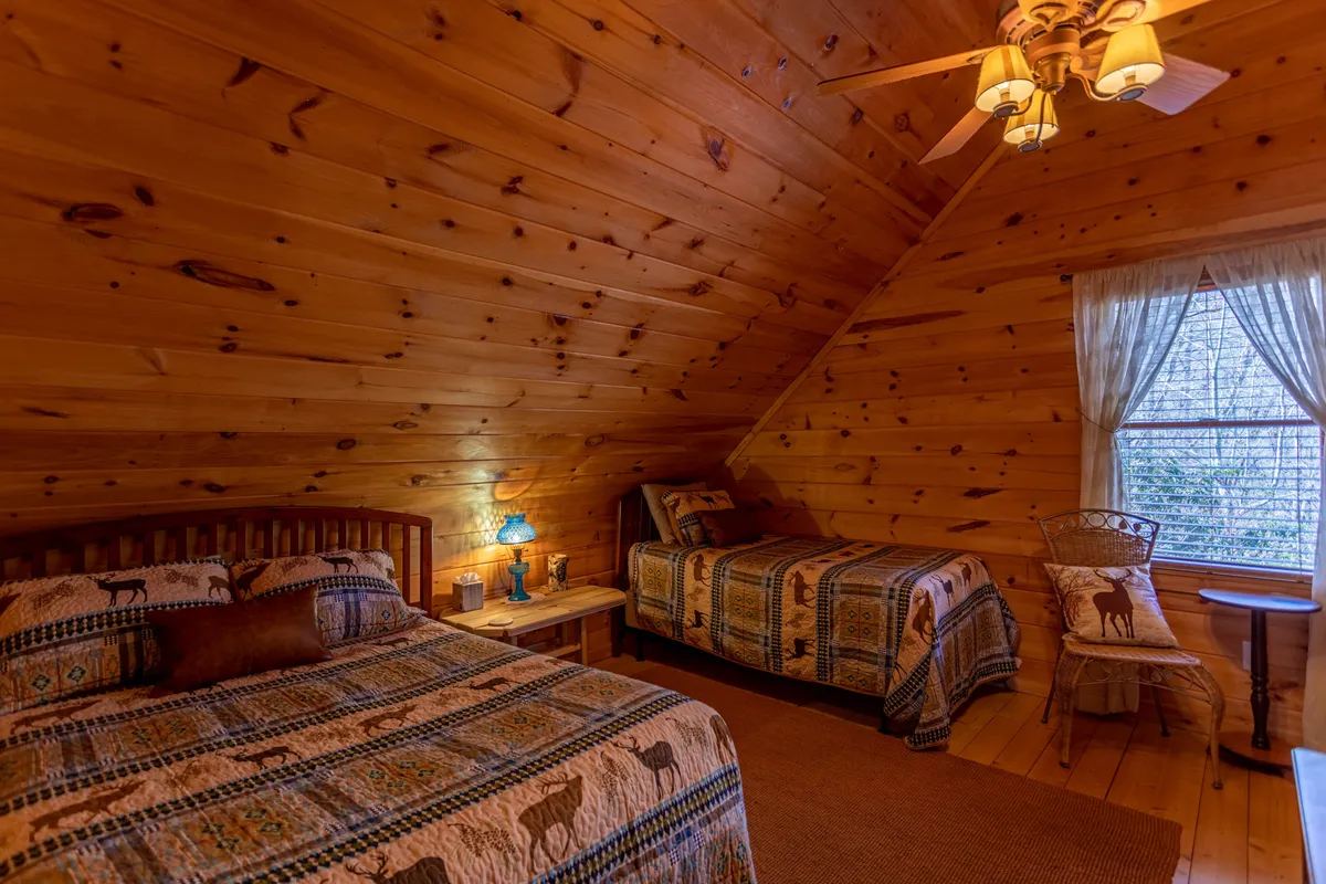 A warmly lit bedroom with wooden walls, patterned bedding, a lamp on the nightstand, and a window offering scenic views. The cozy atmosphere is perfect for reading or relaxing after a day of outdoor activities.