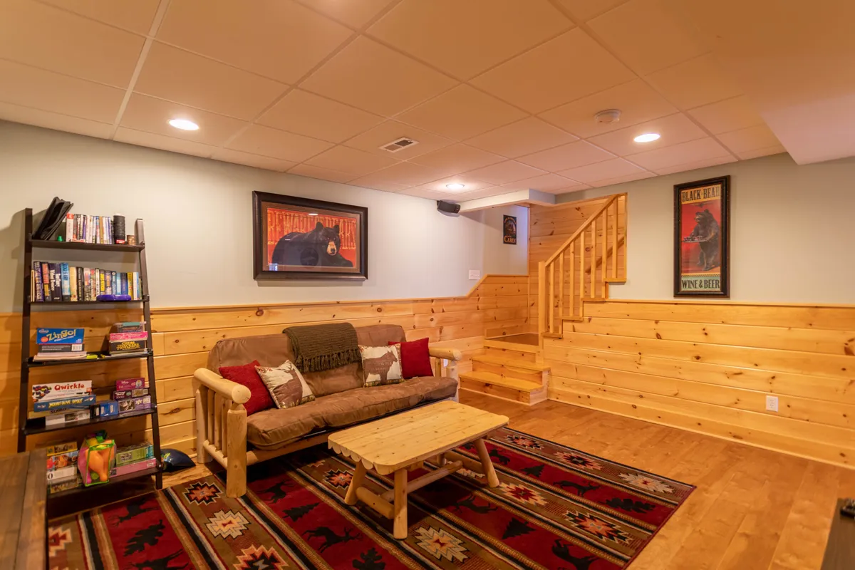 A relaxed lounge space with a plush sofa, wooden coffee table, and a bookshelf stocked with games and movies. The staircase in the background suggests a spacious layout, and the overall mood is casual and welcoming—ideal for movie marathons or game nights.