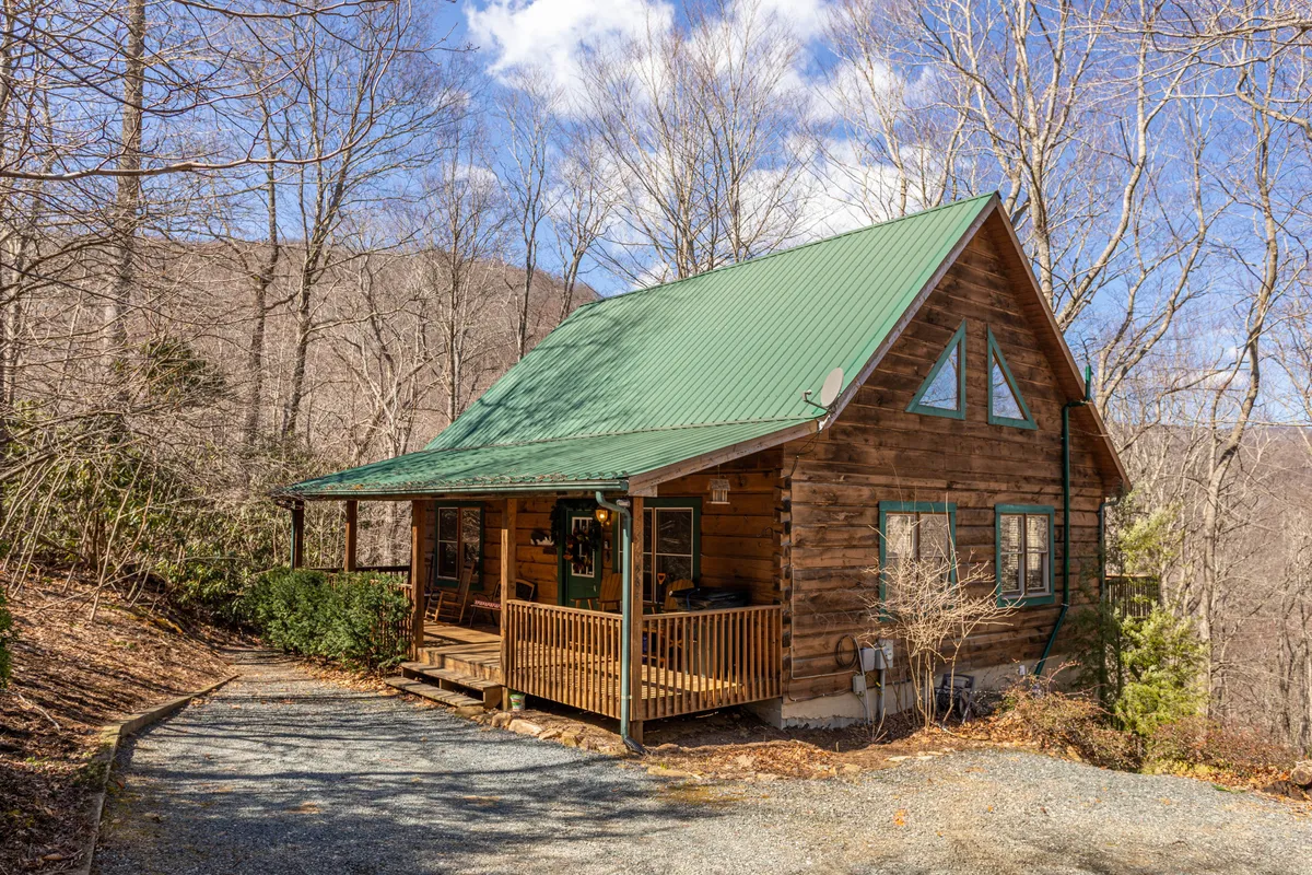 The image shows a charming wooden cabin with a green roof nestled among tall trees. The spacious front porch invites guests to unwind and take in the peaceful, wooded mountain surroundings. The setting is serene, embodying the perfect mountain escape.