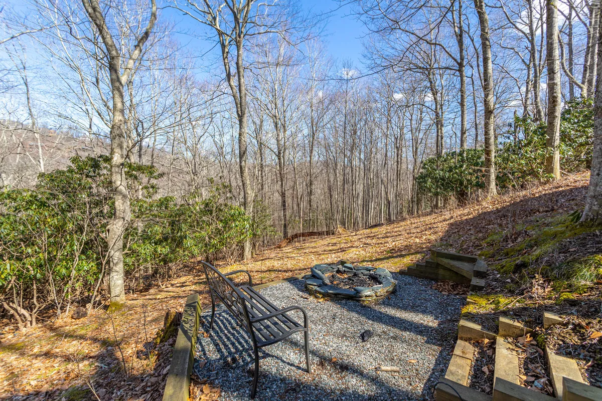 The image depicts a fire pit encircled by bench seating under a canopy of mature trees. The relaxed setup encourages gathering around the fire, storytelling, and stargazing in a peaceful wooded setting.